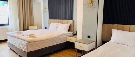 A hotel room with two beds, a large window with curtains, a bedside table with a lamp, and a wall-mounted light fixture.