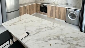 A modern kitchen with a marble countertop, wooden cabinets, a built-in oven, a washing machine, and a stainless steel refrigerator. - Cozy 3 Bedroom Villa Close to the Beach (Limassol)