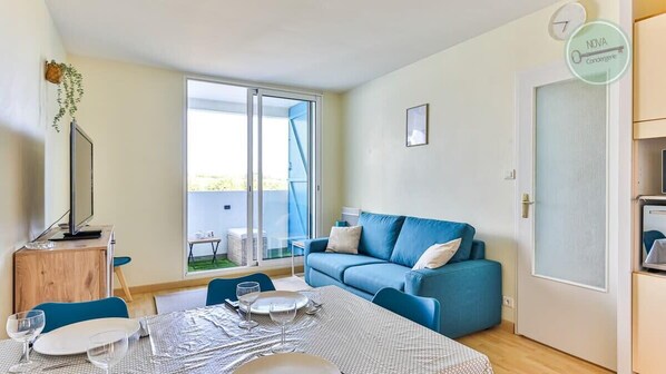 A compact living space with a dining area, a blue sofa, and a sliding glass door leading to a balcony. - La Goëlette 30 Les mouettes (Saint-Hilaire-de-Riez)