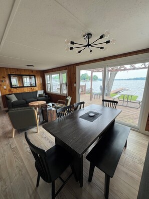 A dining area with a large table and chairs, a wooden floor, and a view of a lake through large windows. - Lakefront 2 Bedroom Cottage Getaway on Chippewa Lake, Michigan (Evart)