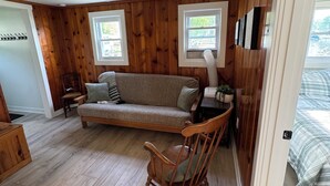Ett mysigt rum med trägolv, en soffa, en gungstol och två fönster med utsikt över grönskan.