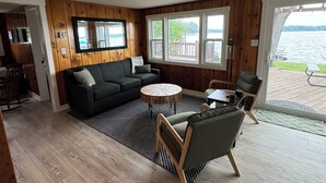 Ett modernt vardagsrum med trägolv, en svart soffa, ett runt soffbord och utsikt över en sjö genom stora fönster.