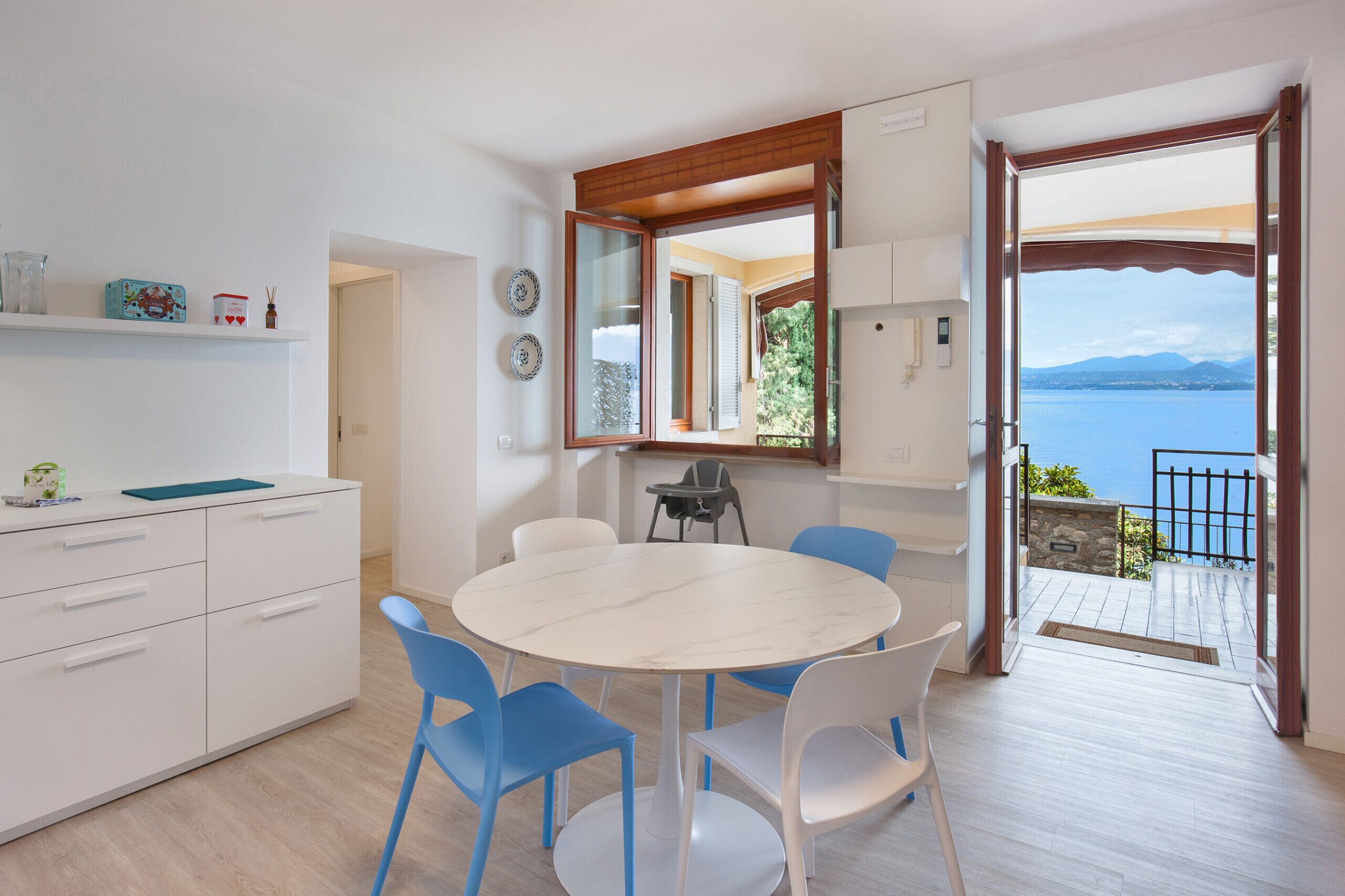 A modern kitchen with white cabinets, a round marble table, and blue chairs. There is a large window with a view of the sea and mountains.