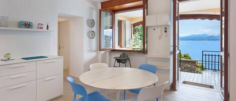 A modern kitchen with white cabinets, a round marble table, and blue chairs. There is a large window with a view of the sea and mountains.