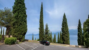 Un aparcamiento con un coche, rodeado de altos cipreses y vistas a un lago y montañas.