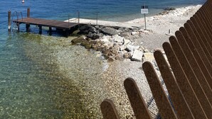 Una vista despejada de una costa rocosa con un muelle de madera que se adentra en aguas tranquilas, un cartel que dice 'Vietato Tuffarsi', y montañas lejanas bajo un cielo claro.