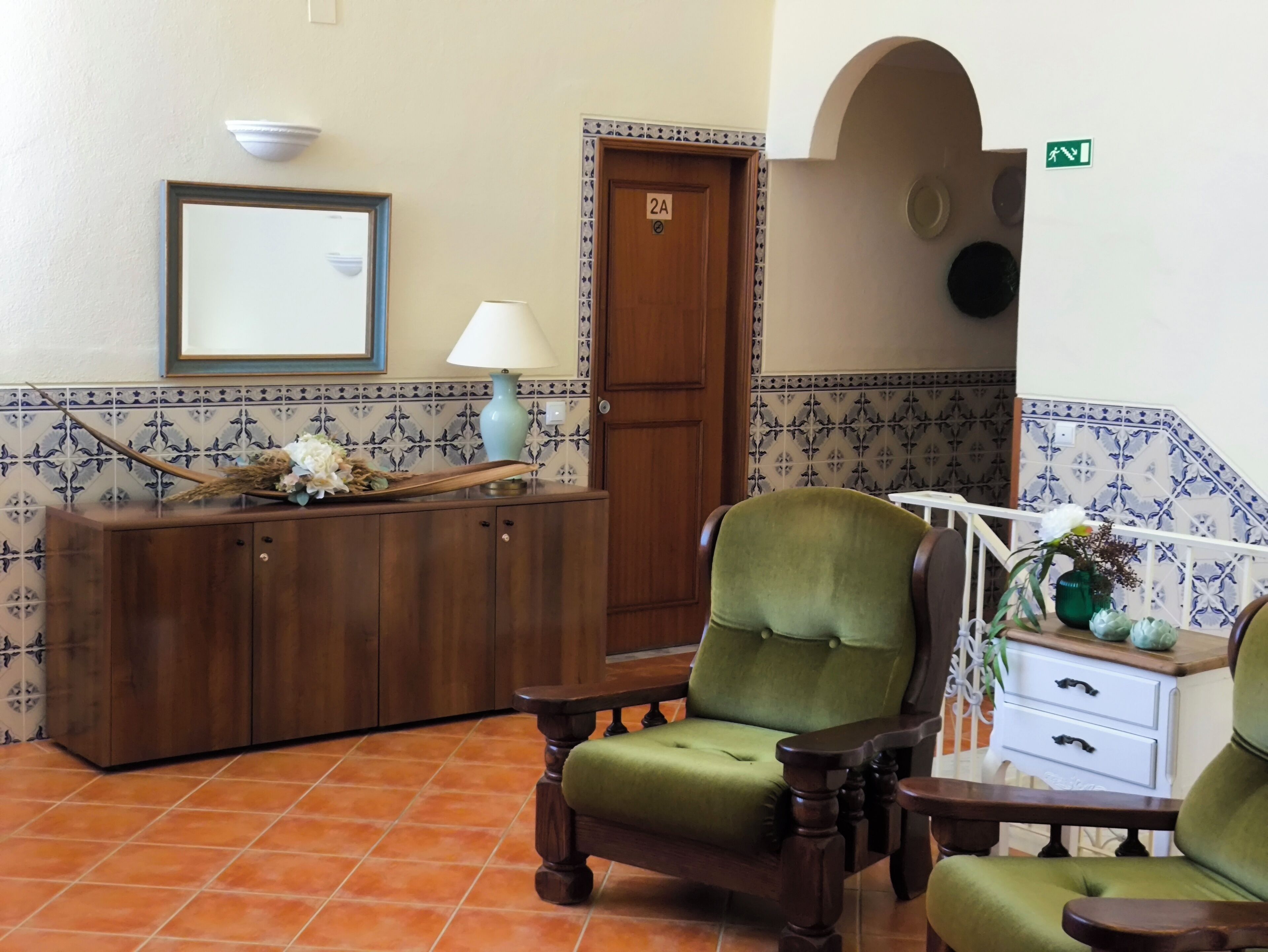 Een hal met een houten receptiebalie, een groene fauteuil, een witte bijzettafel met een vaas, en een betegelde muur met een deur waarop '2A' staat.