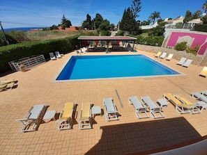 A rectangular swimming pool with a tiled deck, surrounded by white lounge chairs and a covered seating area. - Aurora SOl (Lagos)