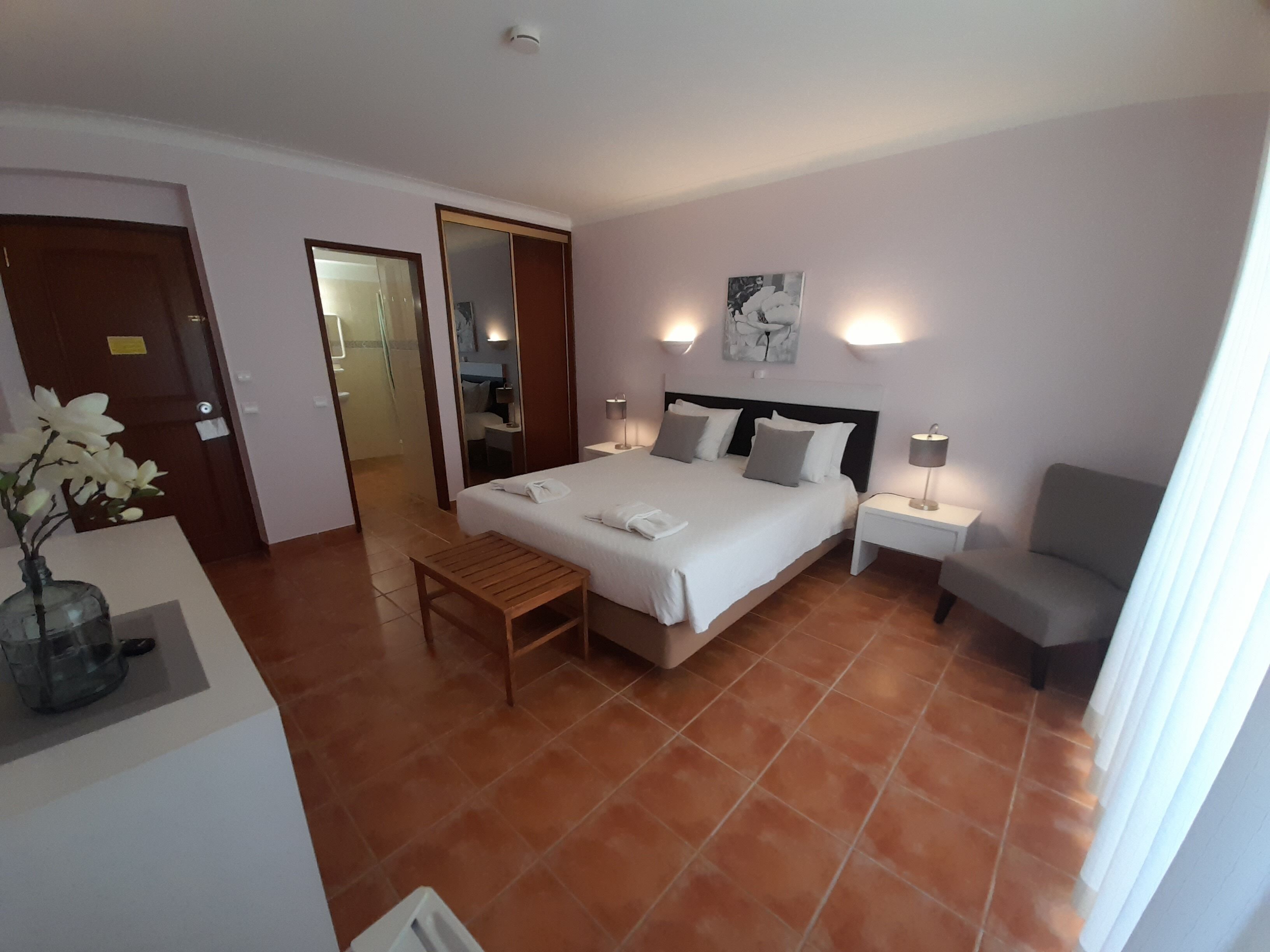 A hotel room with a bed, bedside tables, a chair, a desk with a vase, and a bathroom visible through an open door.