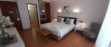 A hotel room with a bed, bedside tables, a chair, a desk with a vase, and a bathroom visible through an open door.