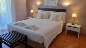 Una cama bien tendida con sábanas y almohadas blancas, dos lámparas de noche, una obra de arte colgada en la pared y un pequeño banco de madera.