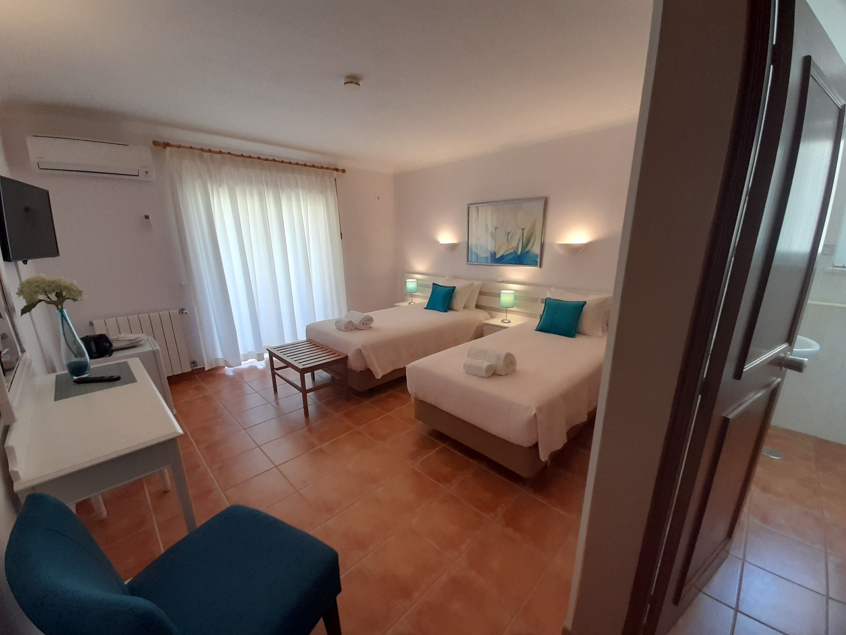 A hotel room with two beds, a desk, a chair, a radiator, and a painting on the wall.