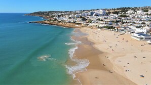 Una playa costera con arena, aguas azules transparentes y una zona residencial con edificios blancos al fondo.