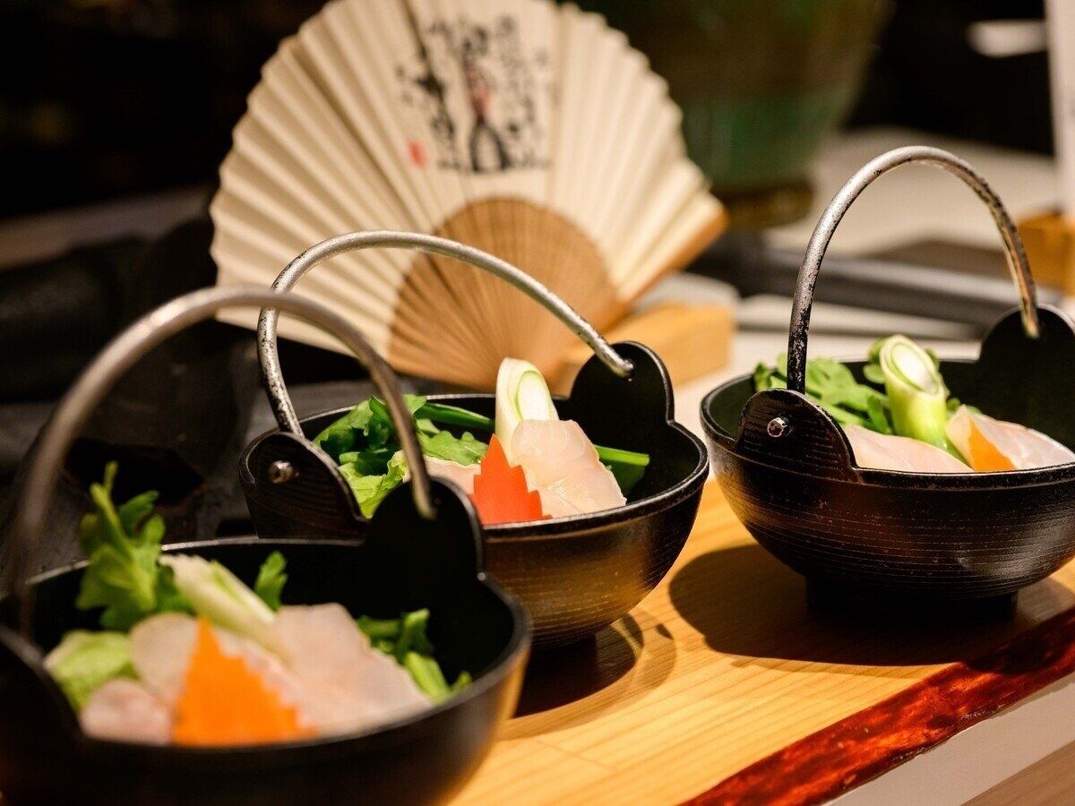 Three bowls of sashimi on a wooden table with a fan in the background.