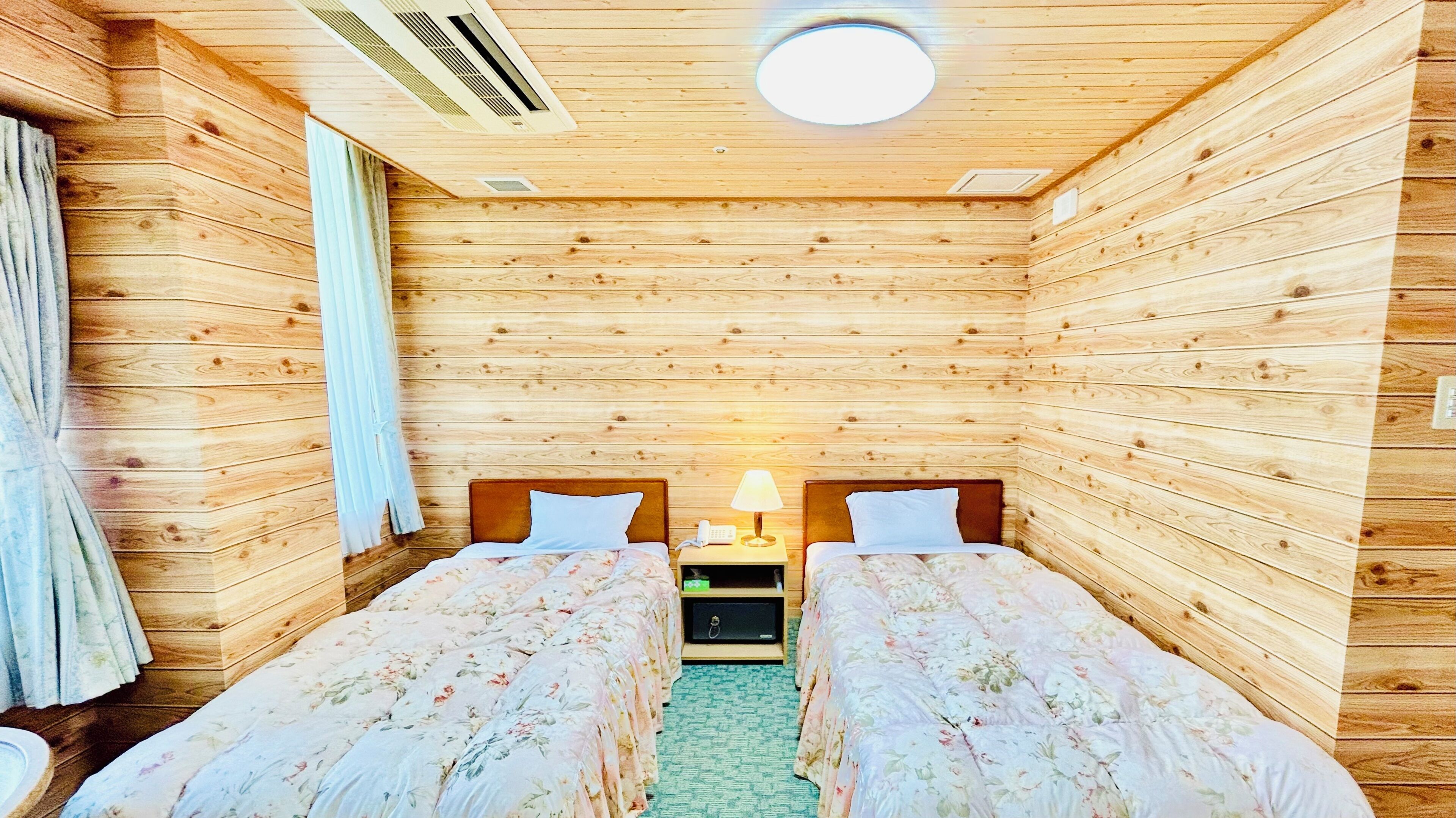 A wooden cabin-style room with two beds, a nightstand with a lamp, and a ceiling vent.
