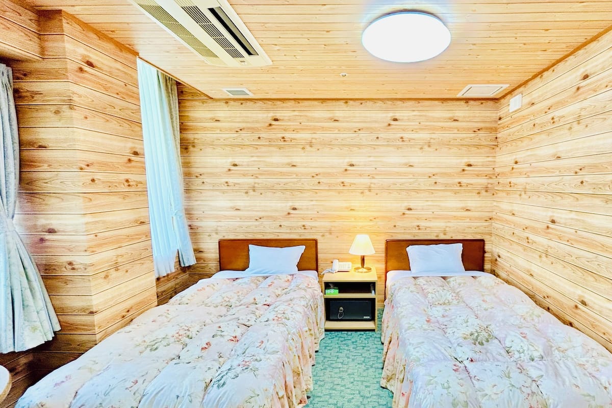 Une pièce aménagée comme un chalet en bois, avec deux lits, une table de chevet avec une lampe et une bouche d’aération au plafond.