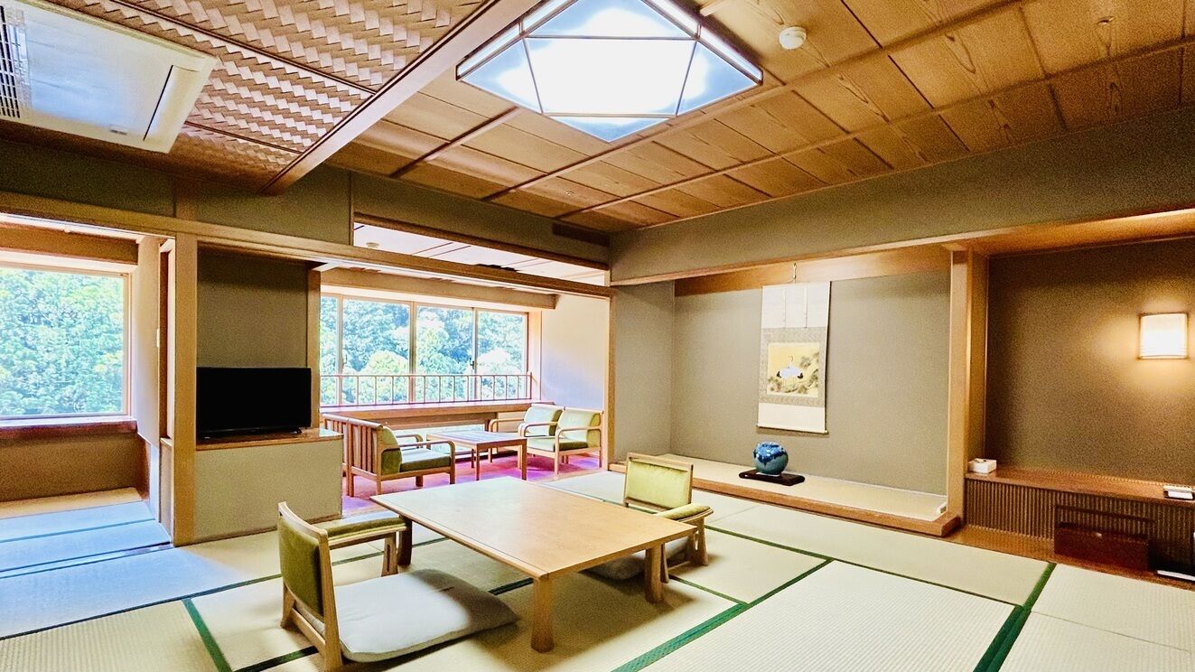 A traditional Japanese room with tatami flooring, a low table, chairs, a television, and a large window with a view of greenery.