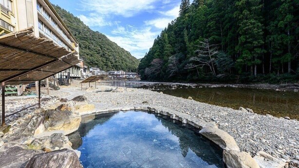 A traditional Japanese onsen with a stone bath surrounded by a rocky landscape and a forested hillside. - Sansuikan Kawayu Midoriya (Tanabe)