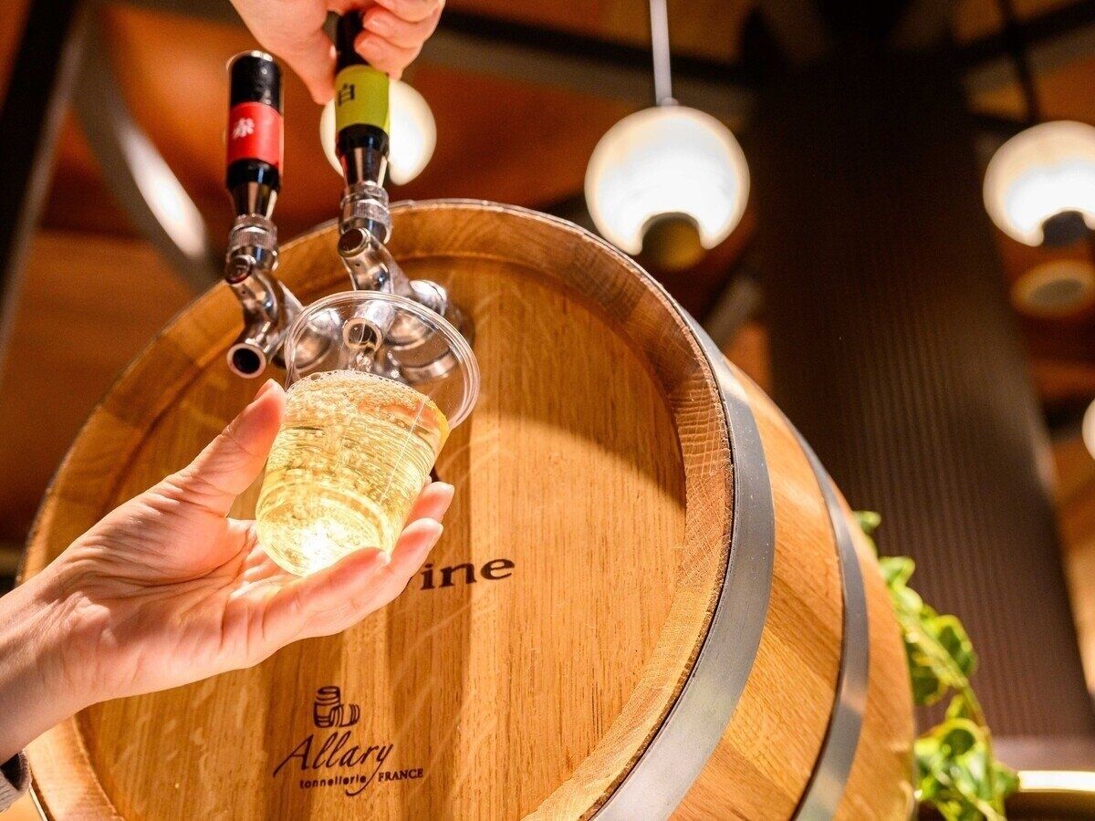 A hand holding a glass of wine with a tap attached, in front of a wooden barrel with the text "Allary" and "France" visible.