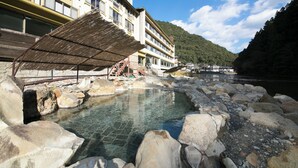A traditional Japanese onsen with a stone bath surrounded by rocks, a wooden walkway, and a building with multiple windows in the background. - Sansuikan Kawayu Midoriya (Tanabe)