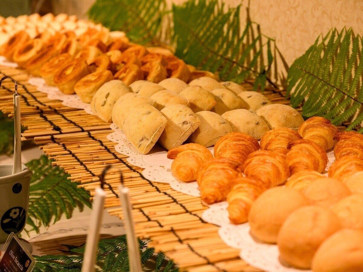 Une table de buffet présentant divers produits de boulangerie, notamment des pâtisseries et des petits pains, disposés sur une surface en bois recouverte d’une nappe en dentelle.