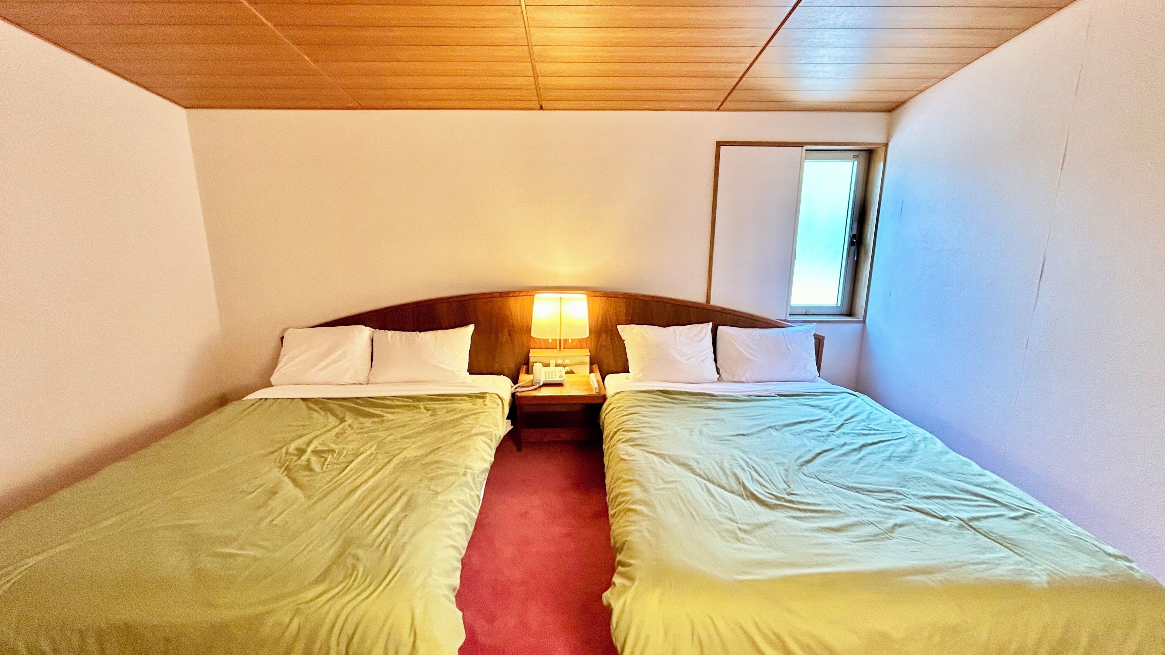 A small hotel room with two single beds, a wooden ceiling, a nightstand with a lamp, and a window.