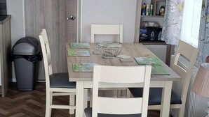 A dining area with a wooden table, white chairs, a black trash bin, and a window with floral curtains. - Beautiful 3-bed Lodge in Kent (New Romney)