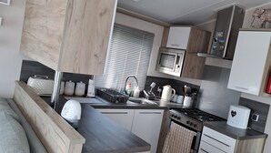 A compact kitchen with a stove, sink, and microwave. There is a window with blinds, a wooden cabinet, and a small refrigerator. - Beautiful 3-bed Lodge in Kent (New Romney)