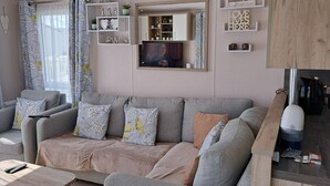 A living room with a grey sofa, floral cushions, a coffee table, and a TV. There are shelves with decorative items and a mirror above the sofa. - Beautiful 3-bed Lodge in Kent (New Romney)