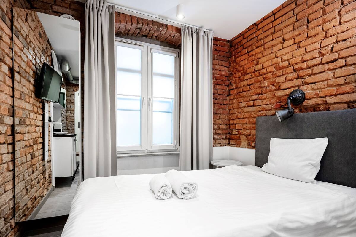 A hotel room with a brick wall, a bed with white linens, a TV mounted on the wall, and a window with curtains.
