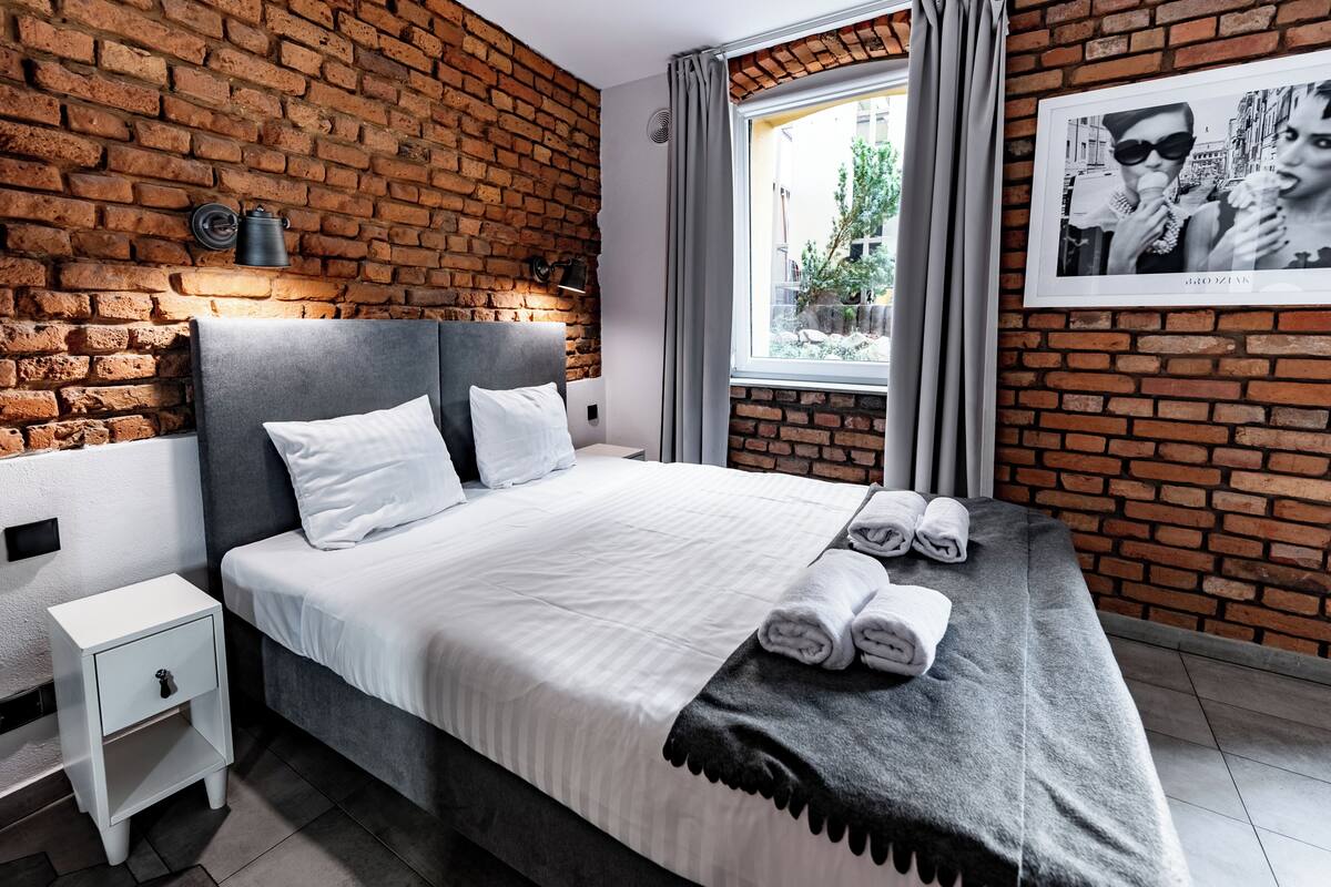 A modern hotel room with a brick wall, a large bed with white linens, a grey headboard, a bedside table with a lamp, and a window with curtains.