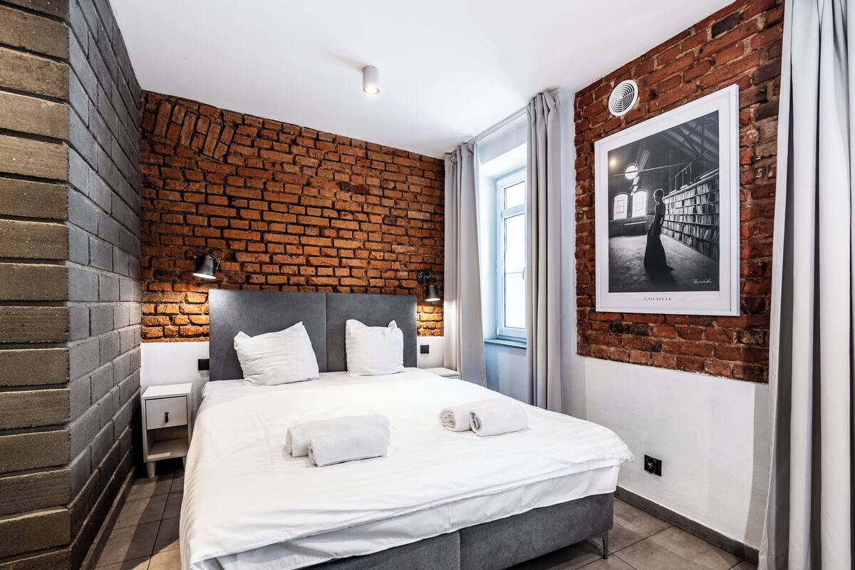 A modern bedroom with a brick wall, a large bed with white bedding, a headboard, and a framed picture on the wall.