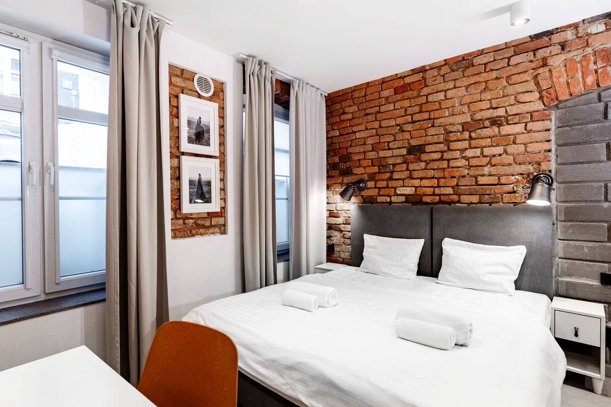 A modern hotel room with a brick accent wall, a neatly made bed, a small bedside table, and a chair.