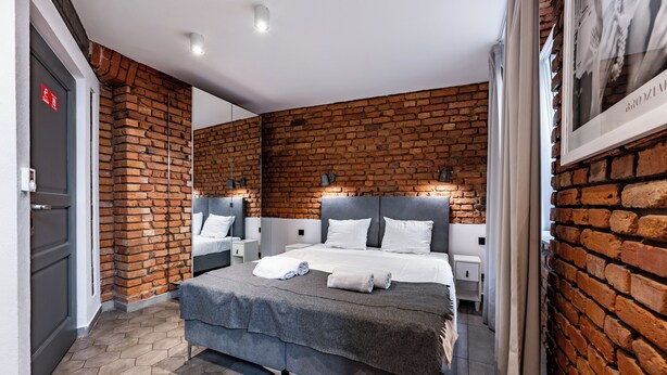 A modern bedroom with a brick wall, a large bed with white bedding, and a small bedside table with a lamp. - RentPlanet - Apartamenty Rybaki (Poznan)