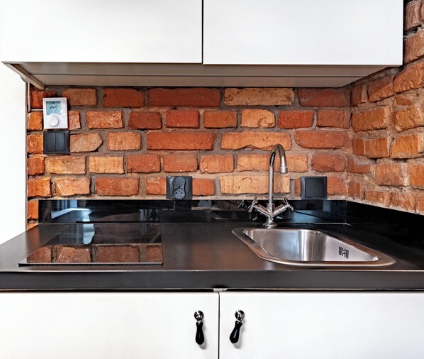 A kitchen with a brick wall backsplash, a stainless steel sink, and white cabinets with black handles. - RentPlanet - Apartamenty Rybaki (Poznan)