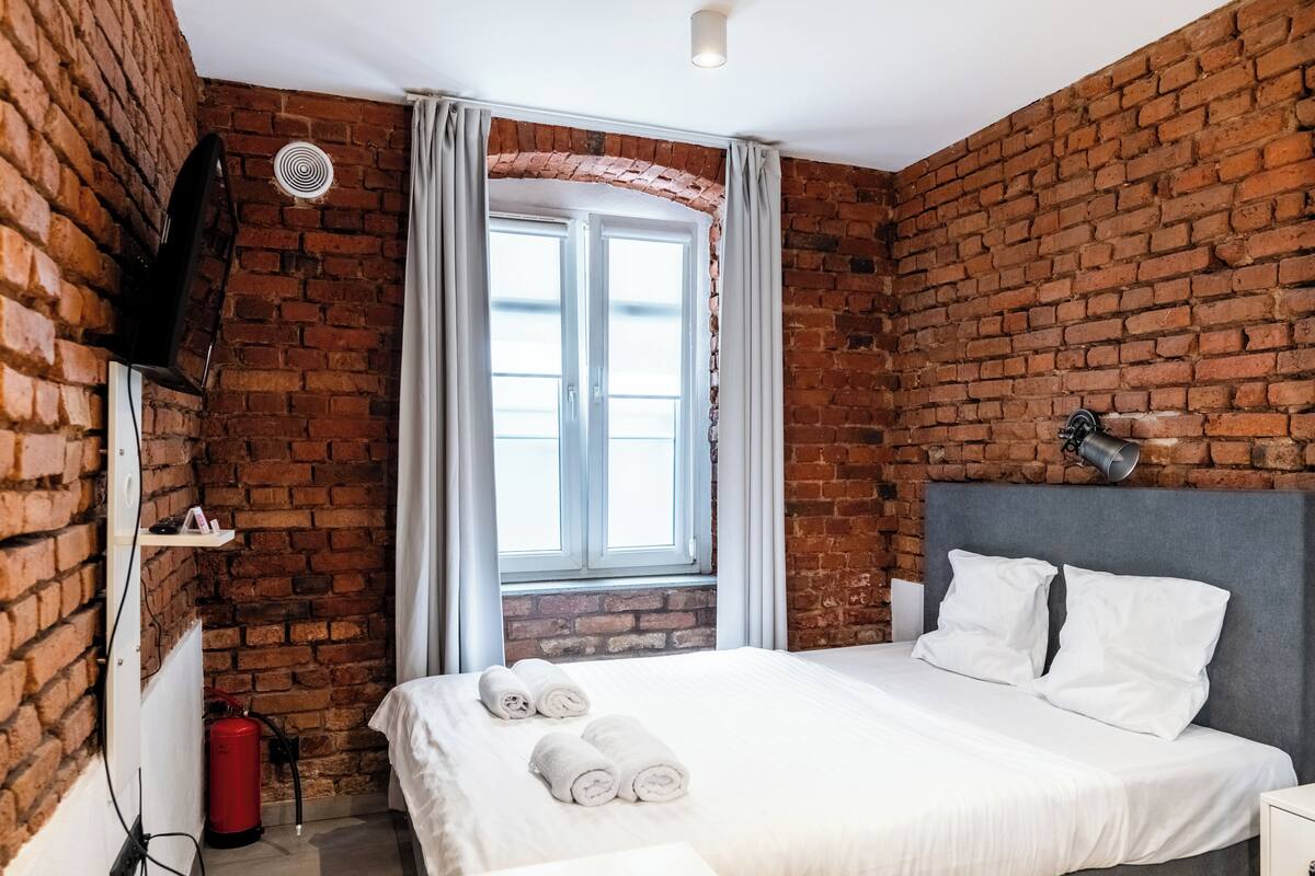 A hotel room with a brick wall, a bed with white bedding, a flat-screen TV mounted on the wall, and a window with curtains.