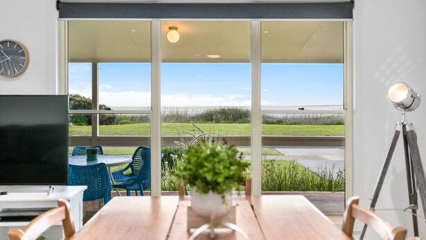 A modern room with a large sliding glass door leading to an outdoor area. Inside, there's a wooden table with chairs, a TV, and a potted plant. - With stunning views, the Blue House is perfect to relax in affordable Luxury.  (Meningie)