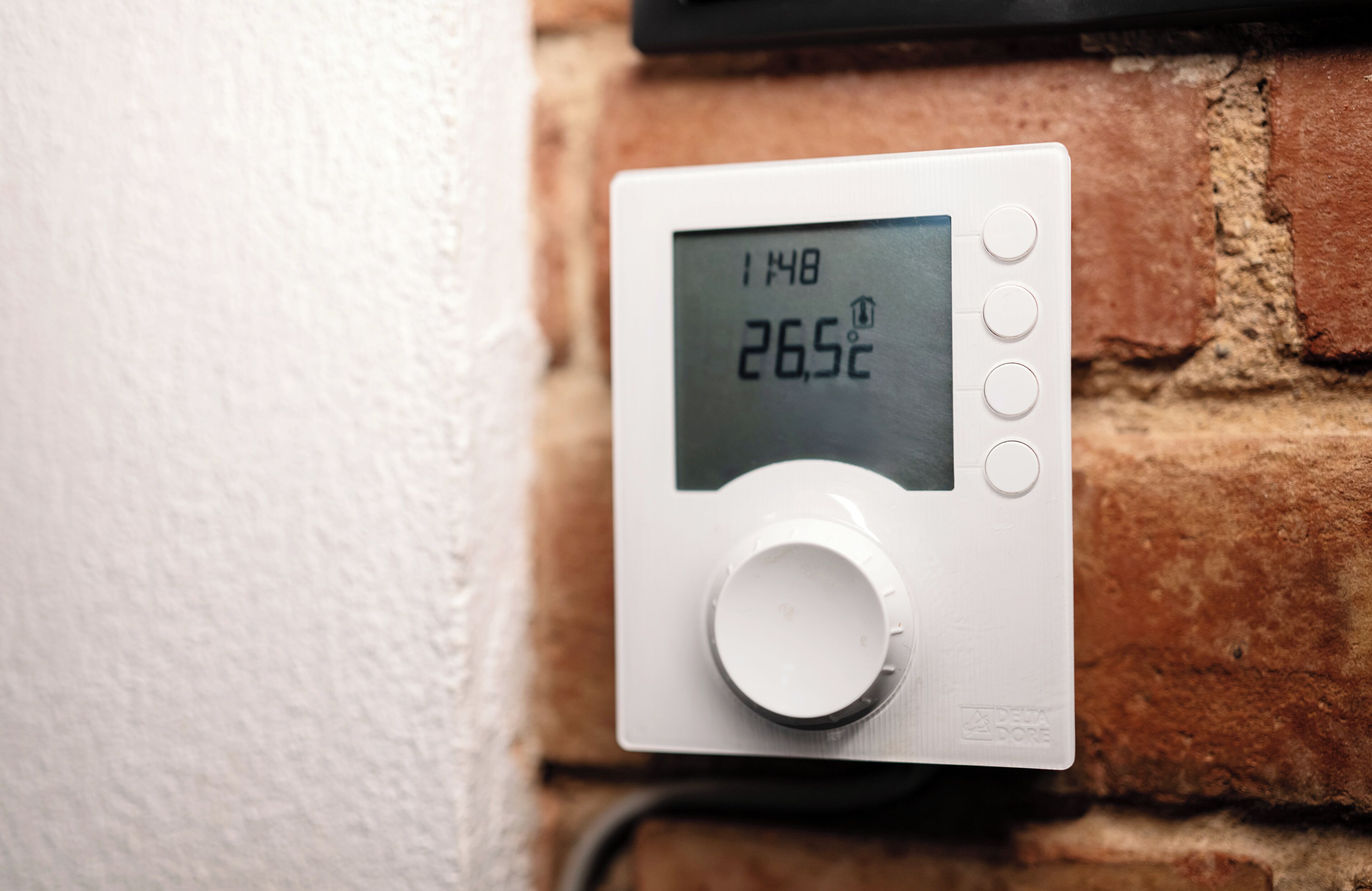 A digital thermostat mounted on a brick wall, displaying a temperature of 26.5°C.