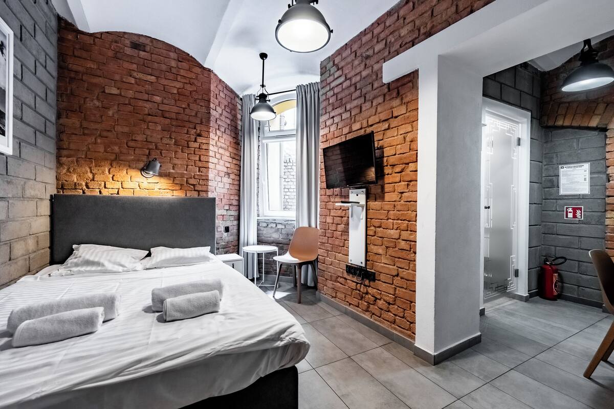 A modern hotel room with a brick wall, a bed with white linens, a small table, and a chair.