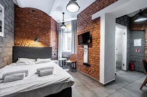Una habitación de hotel moderna con pared de ladrillo, cama con sábanas blancas, una mesita y una silla.