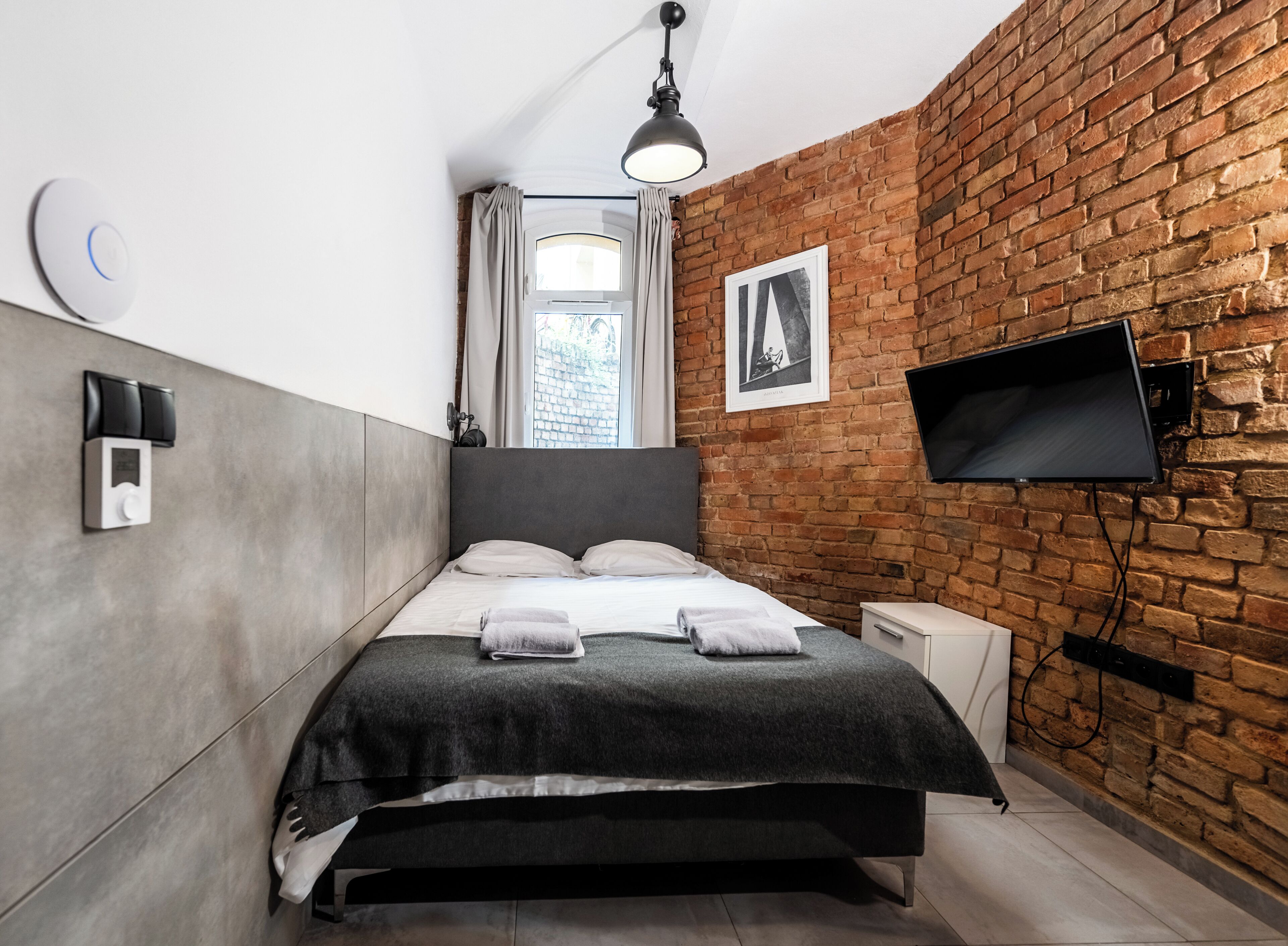 Un dormitorio moderno con pared de ladrillo, cama con cabecera gris, televisor de pantalla plana montado en la pared y ventana con cortinas.