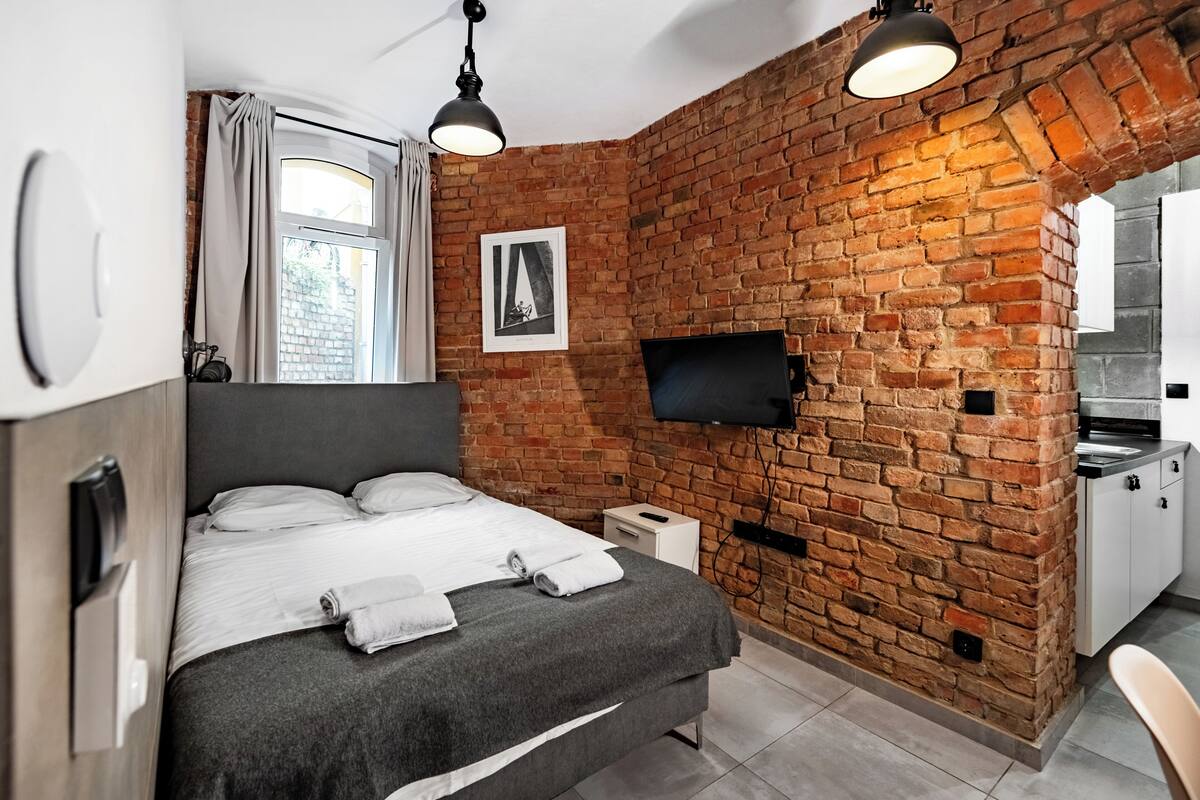 A modern bedroom with a brick wall, a flat-screen TV, a bed with white linens, and a small bedside table.