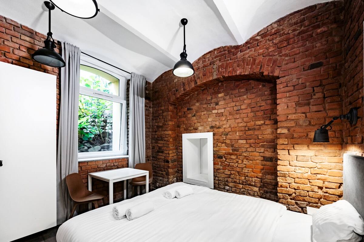 A hotel room with a brick wall, a bed with white linens, a desk with a chair, and a window with curtains.