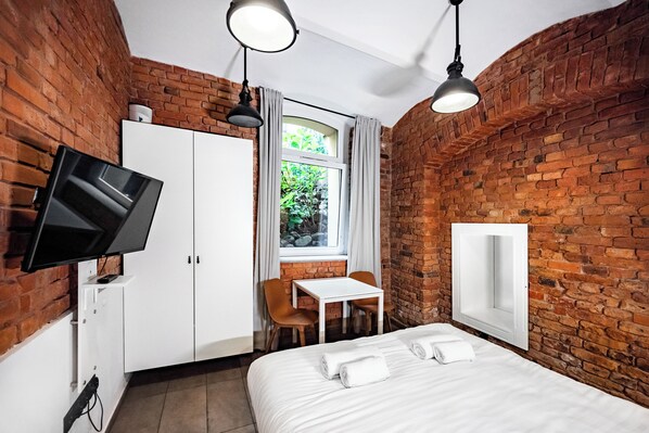 Habitación de hotel con pared de ladrillo, televisor de pantalla plana montado en la pared, cama con ropa de cama blanca, mesita con dos sillas y ventana con cortinas.