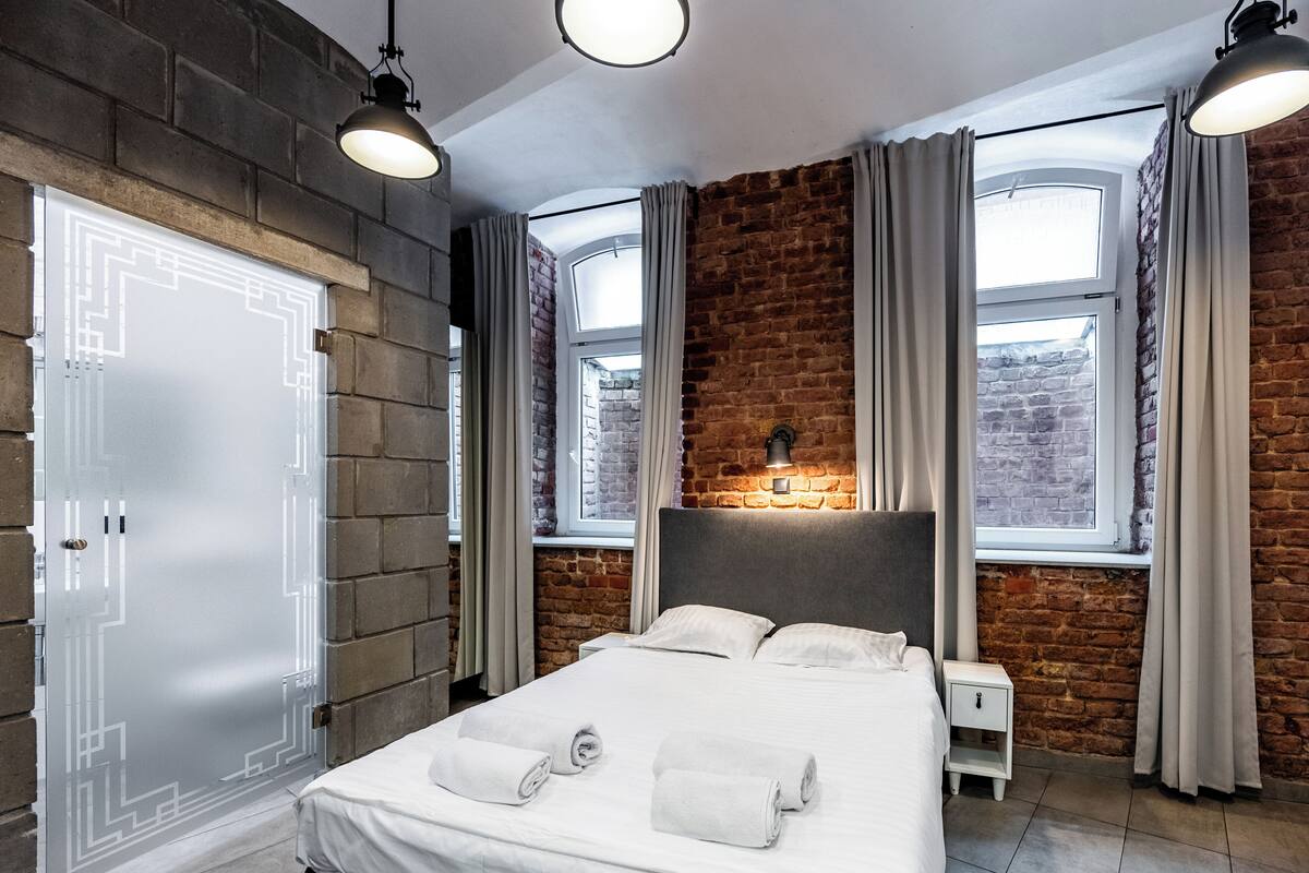 A modern bedroom with a brick wall, a large bed with white linens, two bedside lamps, and a window with curtains.