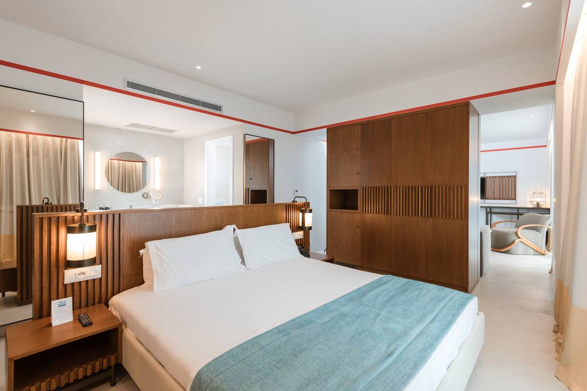 A modern hotel room with a large bed, wooden headboard, a bedside table with a lamp, a mirror, and a wardrobe.