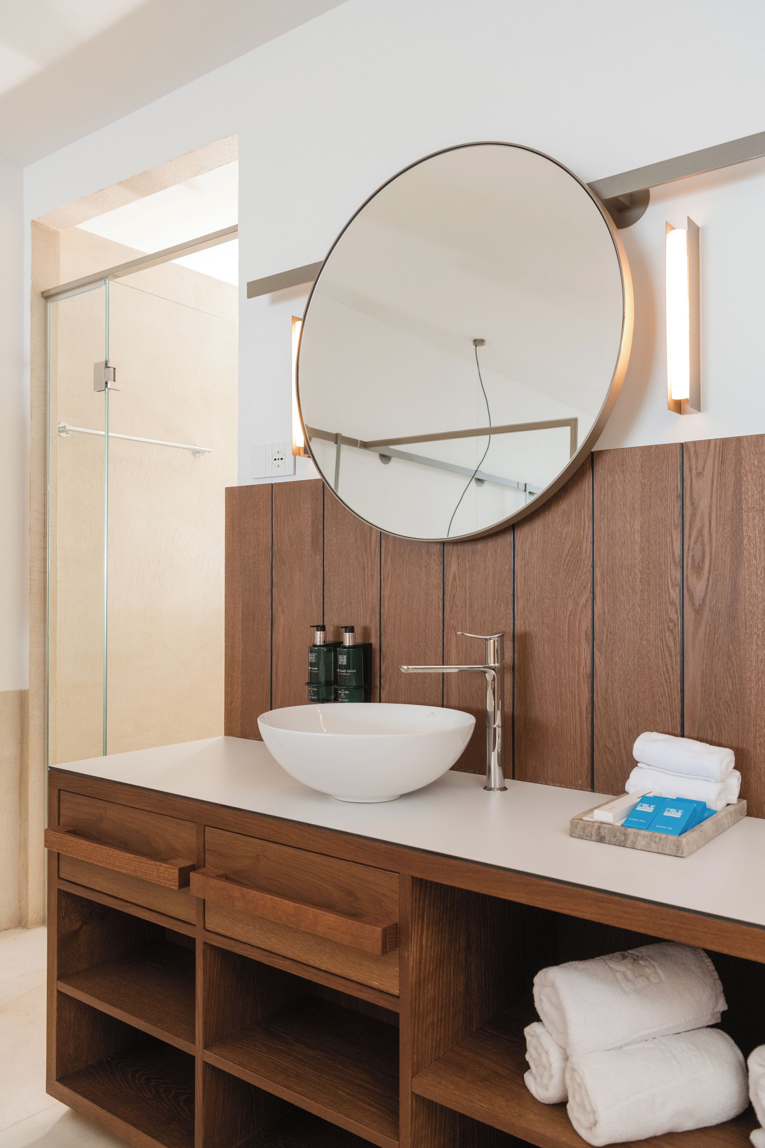 A modern bathroom with a large round mirror, a white sink, wooden cabinetry, and a glass shower enclosure.
