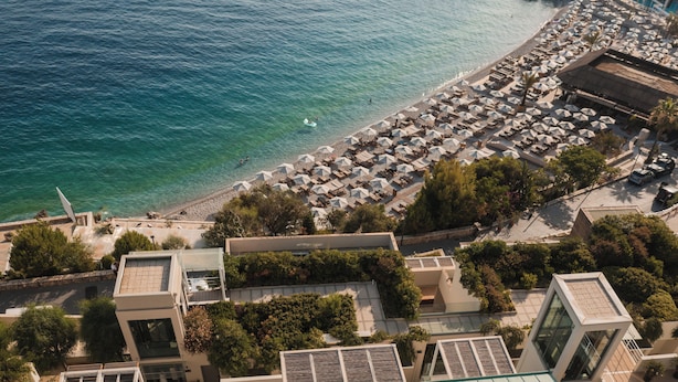 An aerial view of a coastal area with a beach, numerous umbrellas, and buildings. - Folie Marine Beach Hotel & Club (Himare)