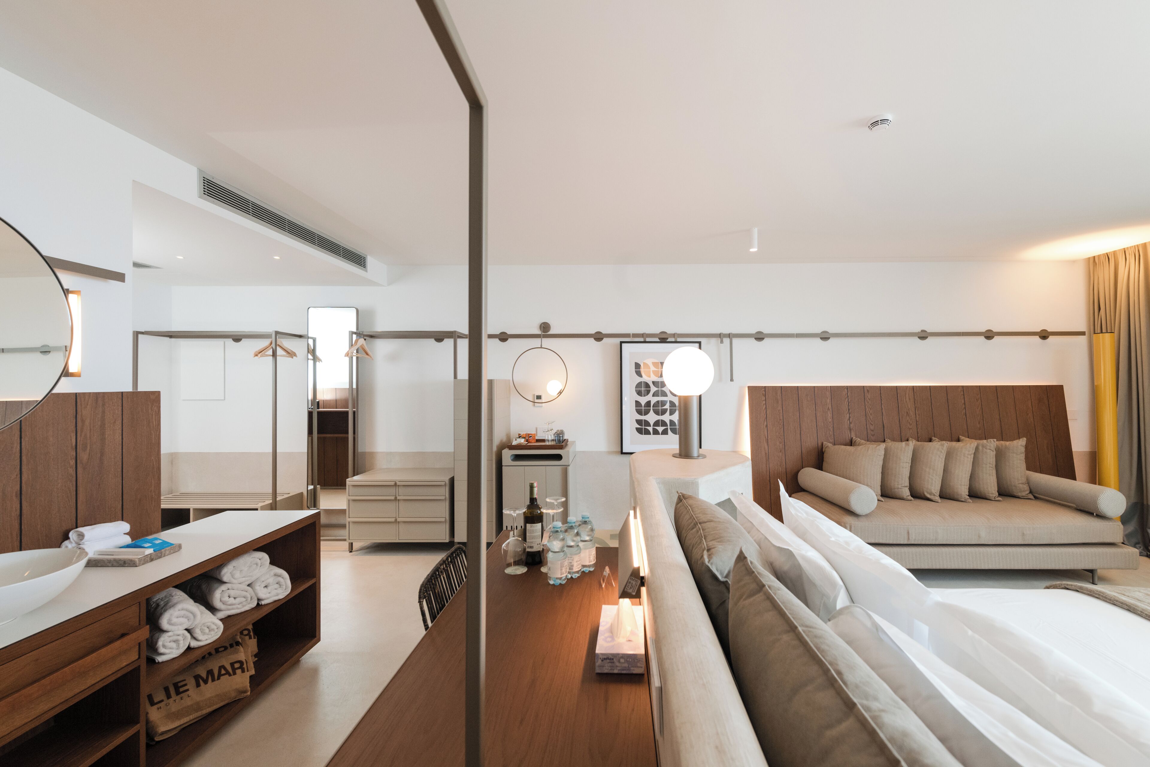 Una habitación de hotel moderna con un tocador de madera, una cama-sofá, una mesa de comedor con un tazón y toallas, y una mesita de noche con botellas y vasos.