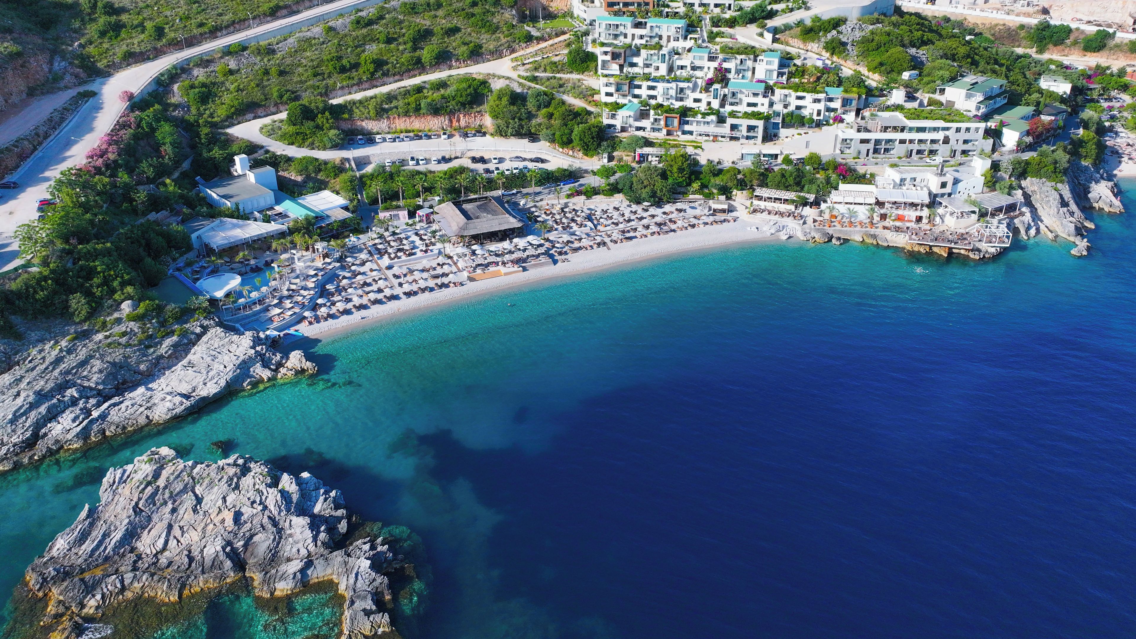 An aerial view of a coastal resort with a beach, parking area, and multiple buildings.